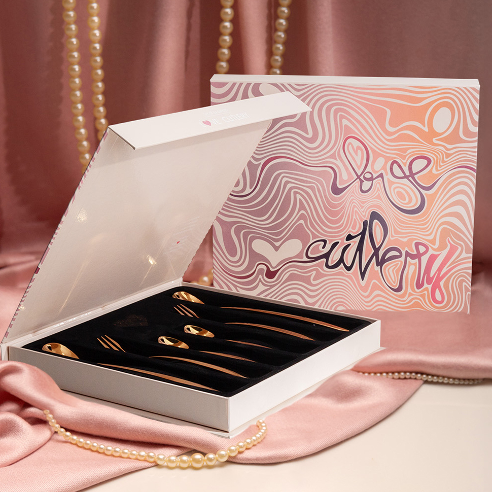 Une boîte blanche ouverte avec des cuillères dorées Love Cutlery, posée sur un satin rose 