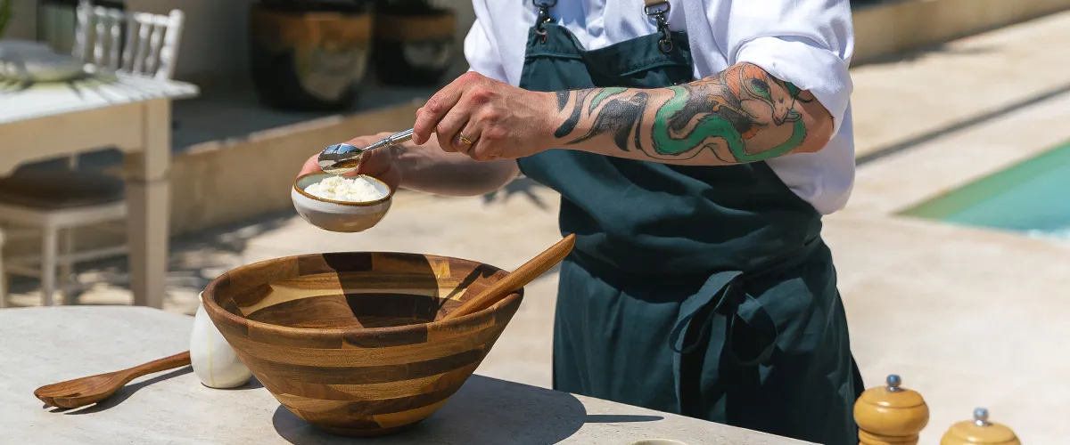 Un chef prépare un plat à côté d’un grand saladier en bois posé sur une table à l’extérieur, près de la piscine 
